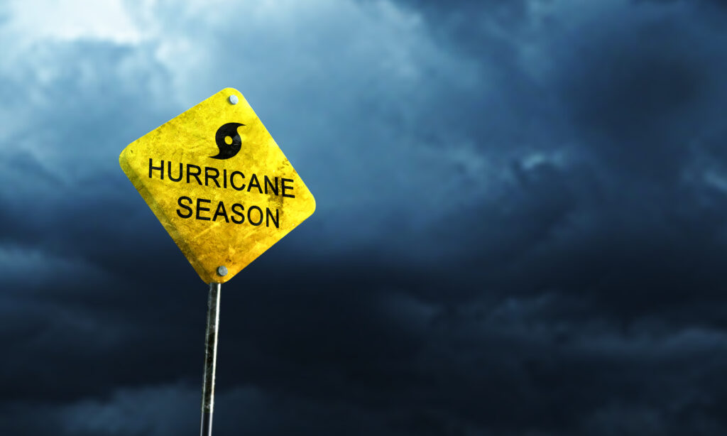 A yellow sign reading "Hurricane Season" stands against a dark, stormy sky, symbolizing the need for understanding hurricane threats and upcoming forecasts.